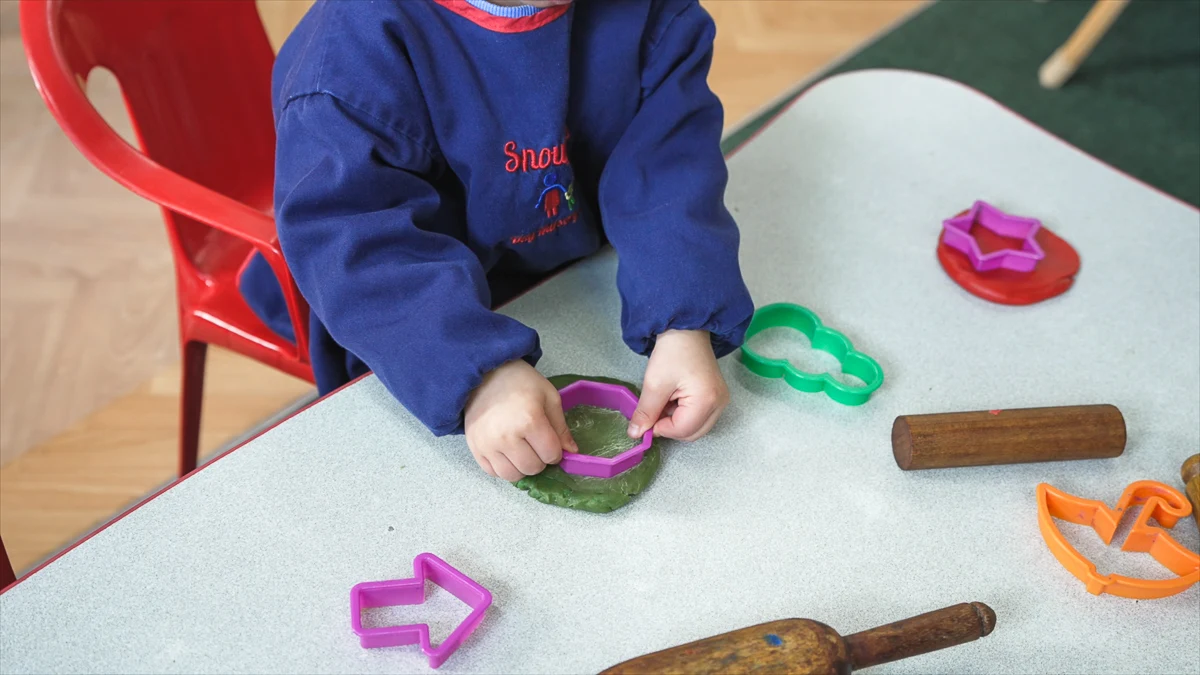 Child pressing a purple shape cutter into green play dough on a table with various other colorful cutters and wooden rolling pins nearby.