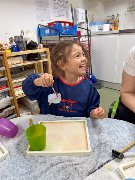 Smiling young girl holding a small craft object during an arts and crafts activity in a classroom setting.
