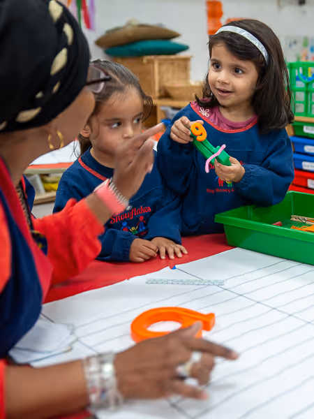 Two young girls interacting with a teacher at a craft table, one girl holding colorful pipe cleaners and buttons.