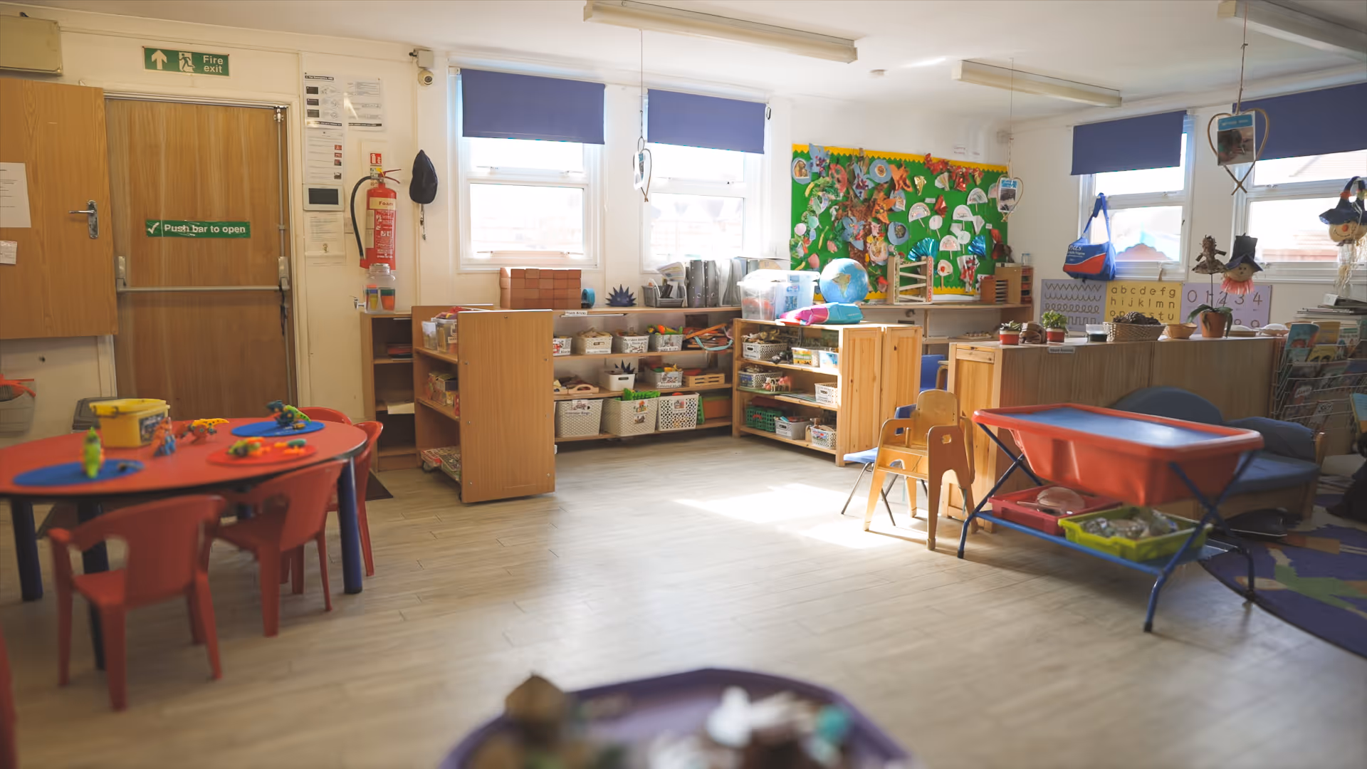 Bright kindergarten classroom with red children’s table and chairs, shelves filled with toys and learning materials, and colorful educational displays on the walls.