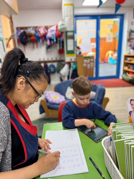 Teacher writing in a notebook while a young boy uses a tablet at a green table in a colorful classroom.