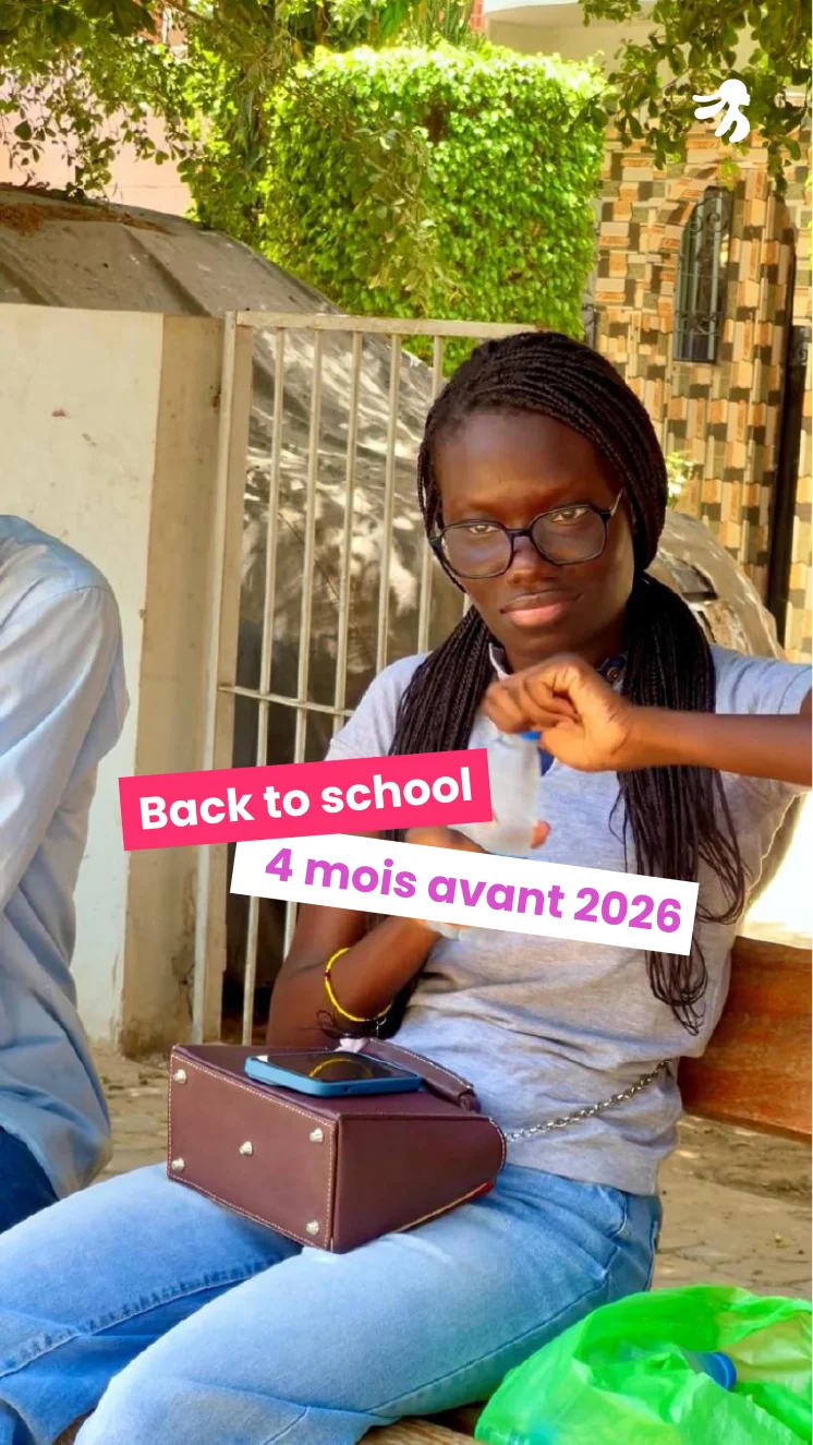 Young woman with glasses sitting outdoors holding a plastic bottle, a brown bag and smartphone on her lap, with green foliage and a building in the background.