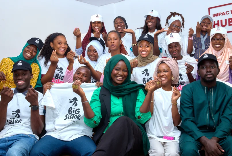 Group of diverse young women and men smiling, wearing Big Sister Africa shirts and caps, posing together indoors.