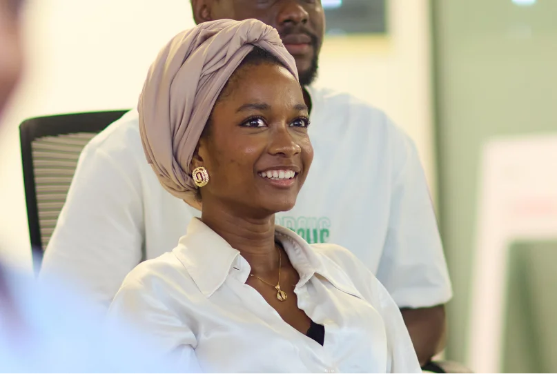 Smiling woman wearing a light-colored headwrap and white blouse sitting indoors with a man in the background.