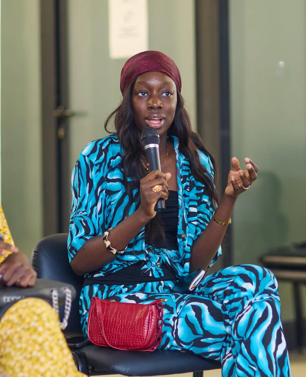 Woman wearing a blue and black patterned outfit and red headscarf speaking into a microphone while seated.