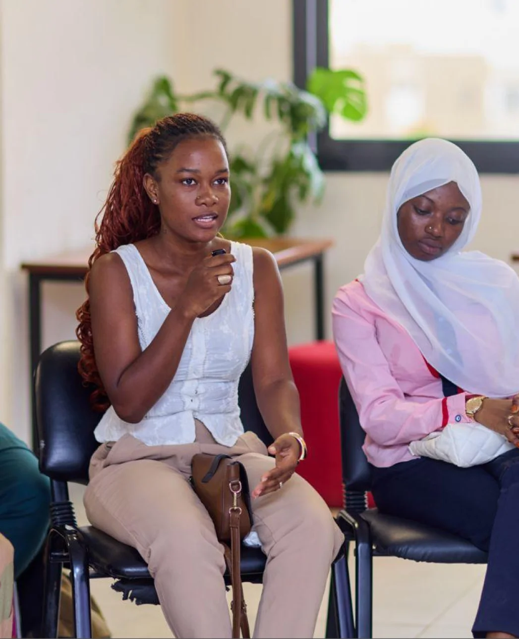 Two women sitting on chairs in a discussion, one speaking while gesturing with her hand, the other listening with her head down wearing a white headscarf.