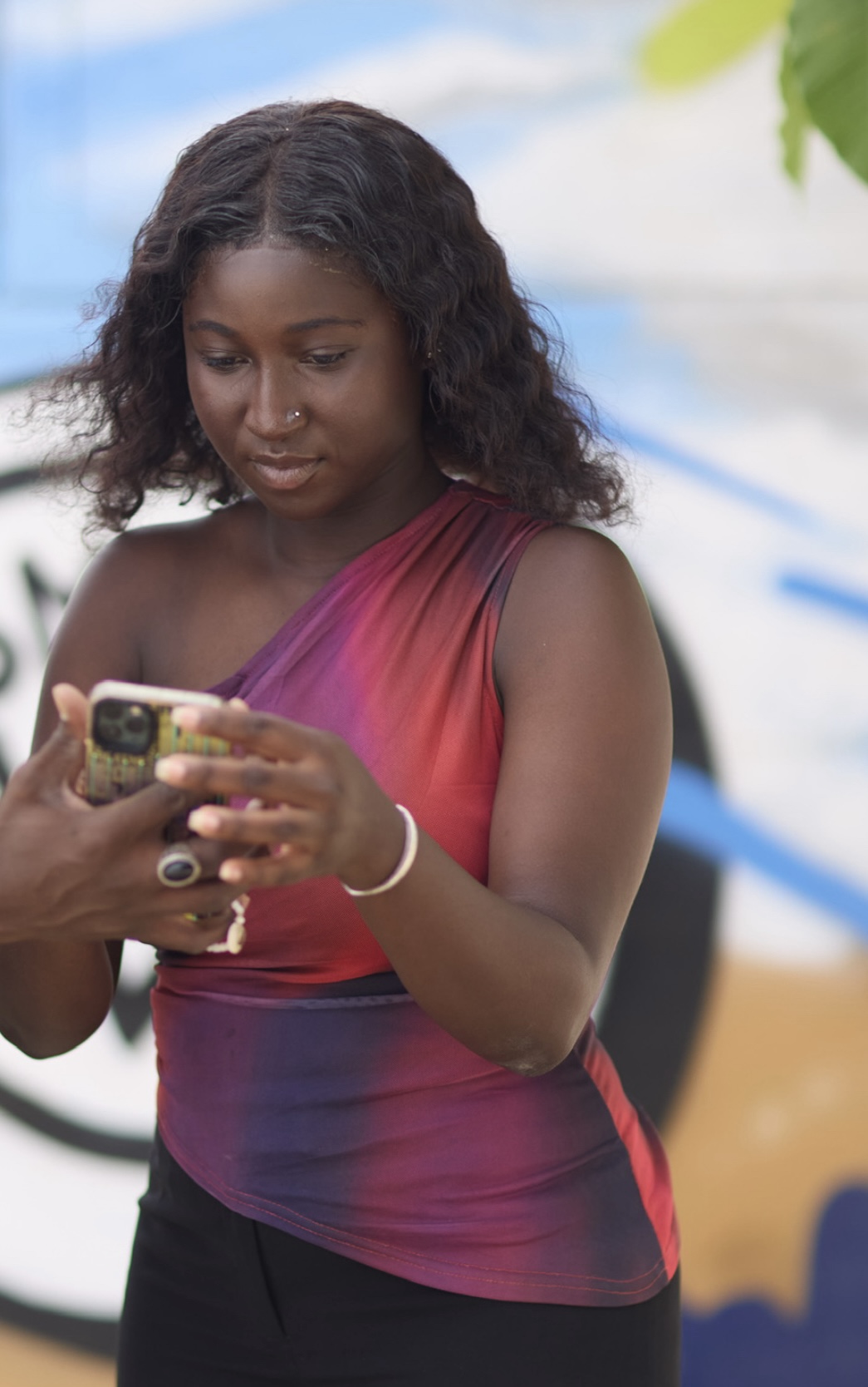 Young woman with curly hair wearing a red and purple sleeveless top looking at a phone held by another person.