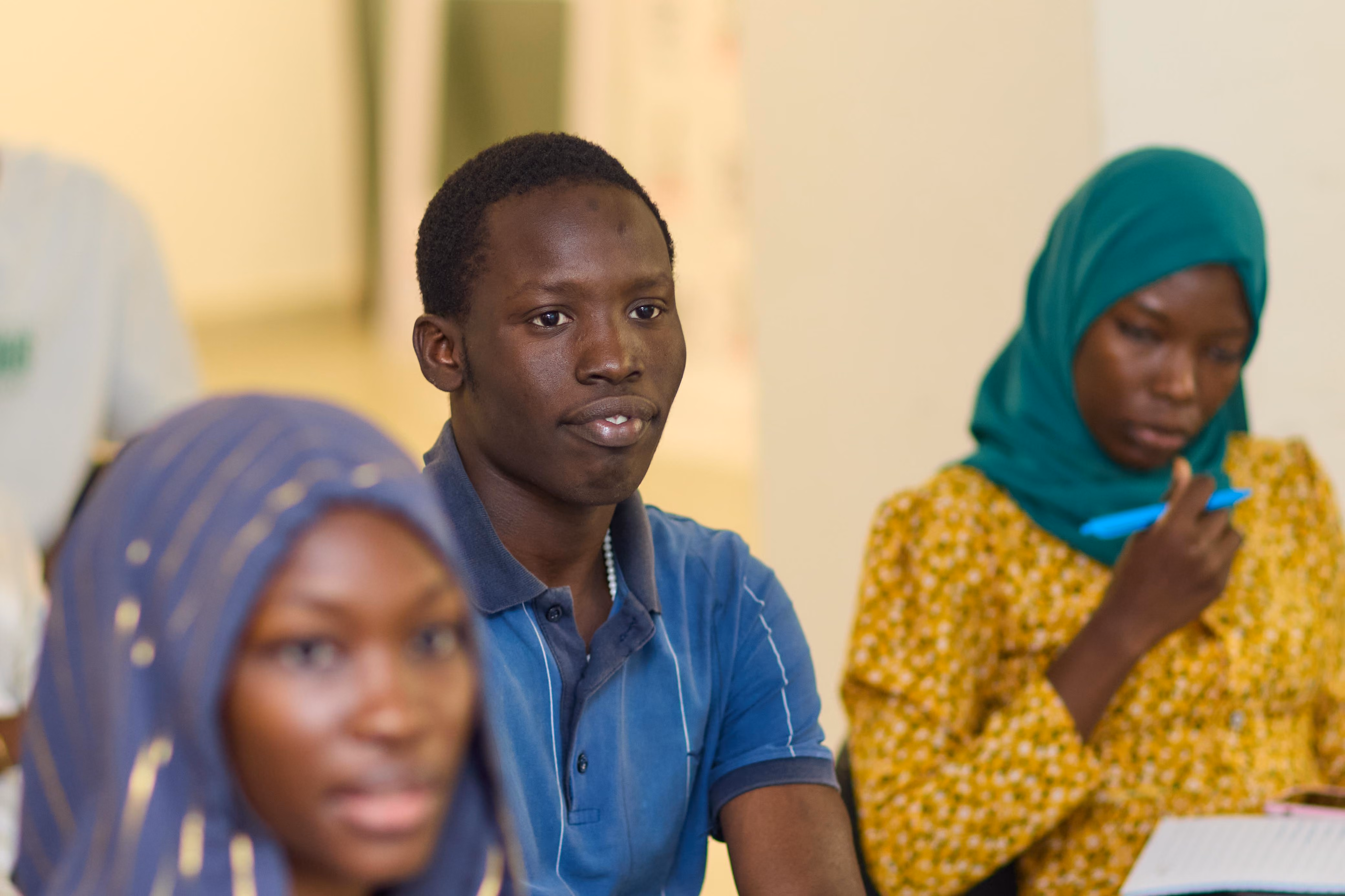 Three young people in a classroom setting, with a young man in a blue shirt in focus and two young women wearing headscarves, one holding a pen and notebook.