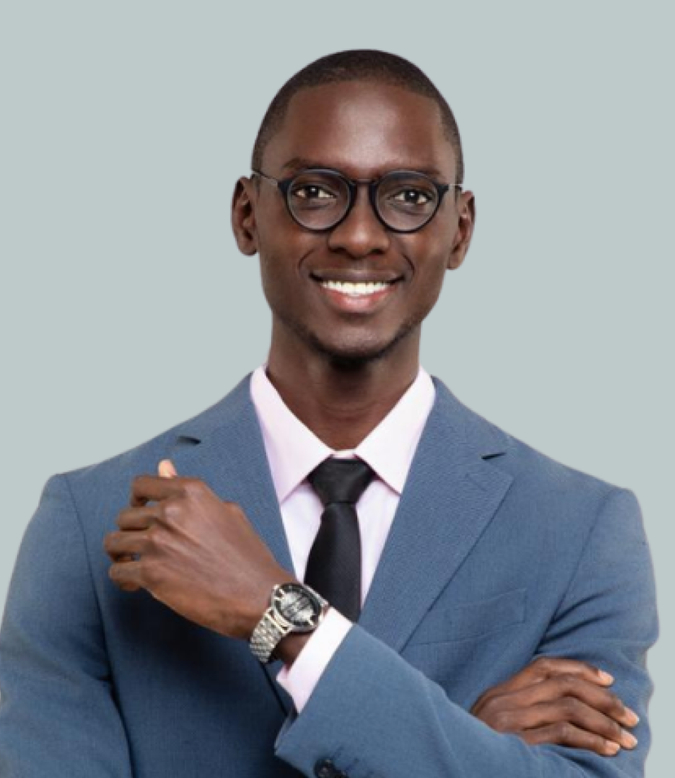 Smiling man wearing glasses, a blue suit, pink shirt, black tie, and a silver wristwatch, standing with arms crossed.