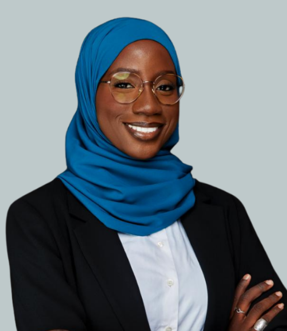 Smiling woman wearing a blue hijab, glasses, white shirt, and black blazer with arms crossed.