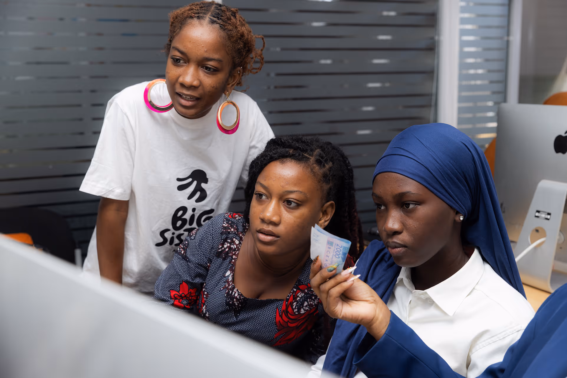 Three young women focused on a computer screen, one holding a 2000 Nigerian naira note.