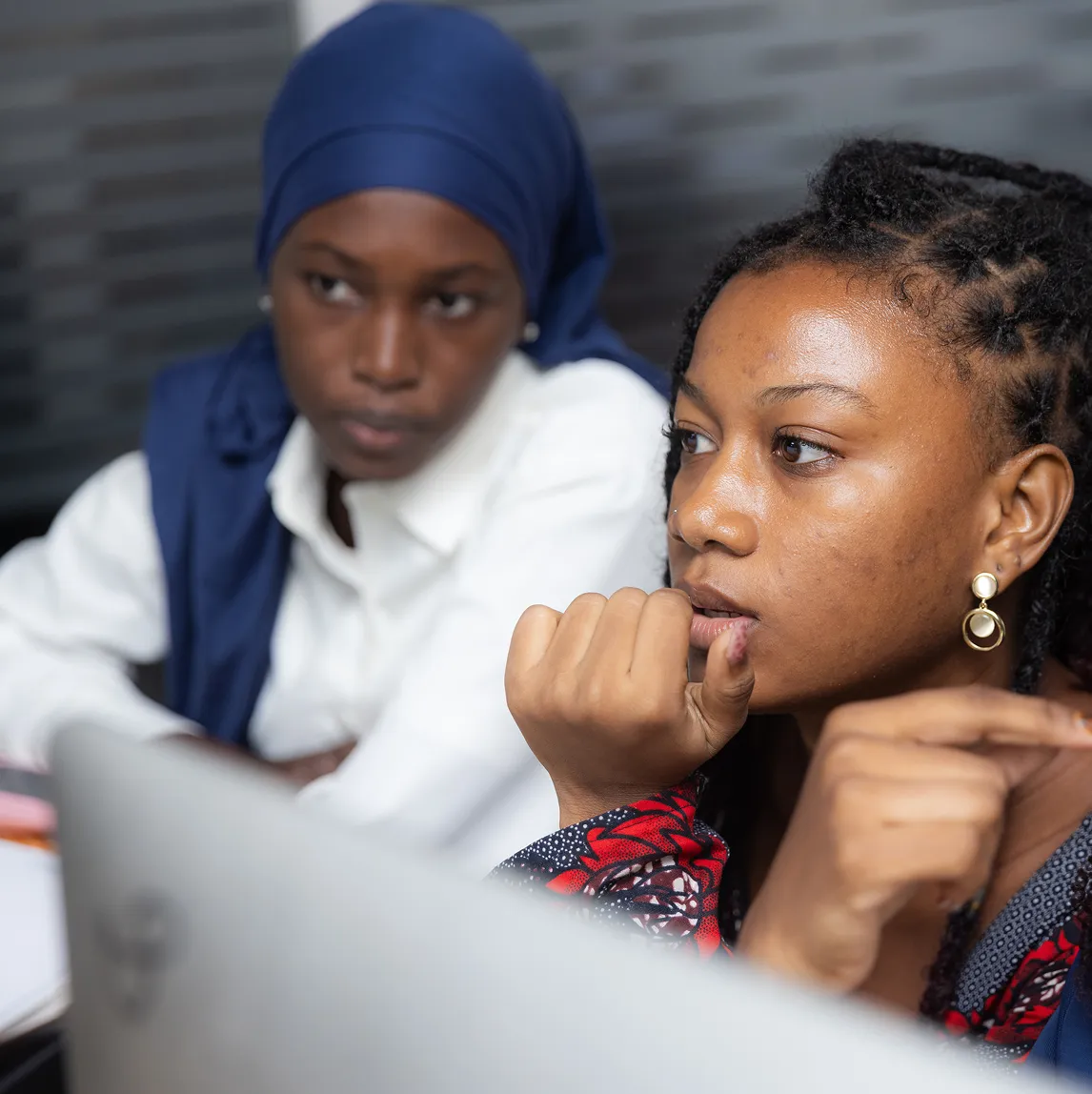 Two women engaged in a thoughtful discussion, one with braided hair wearing earrings, and the other in a blue headscarf and white shirt.