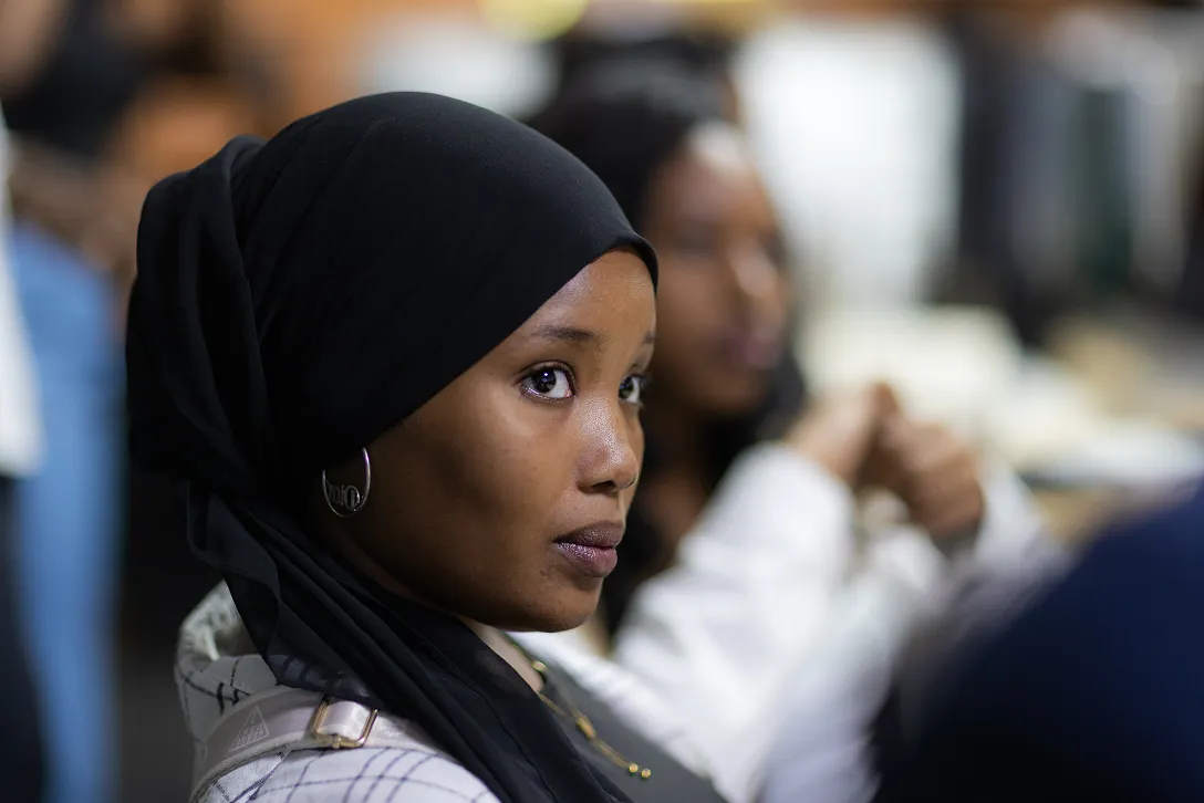 Portrait of a woman wearing a black hijab and silver hoop earrings looking to the side in a softly blurred indoor setting.