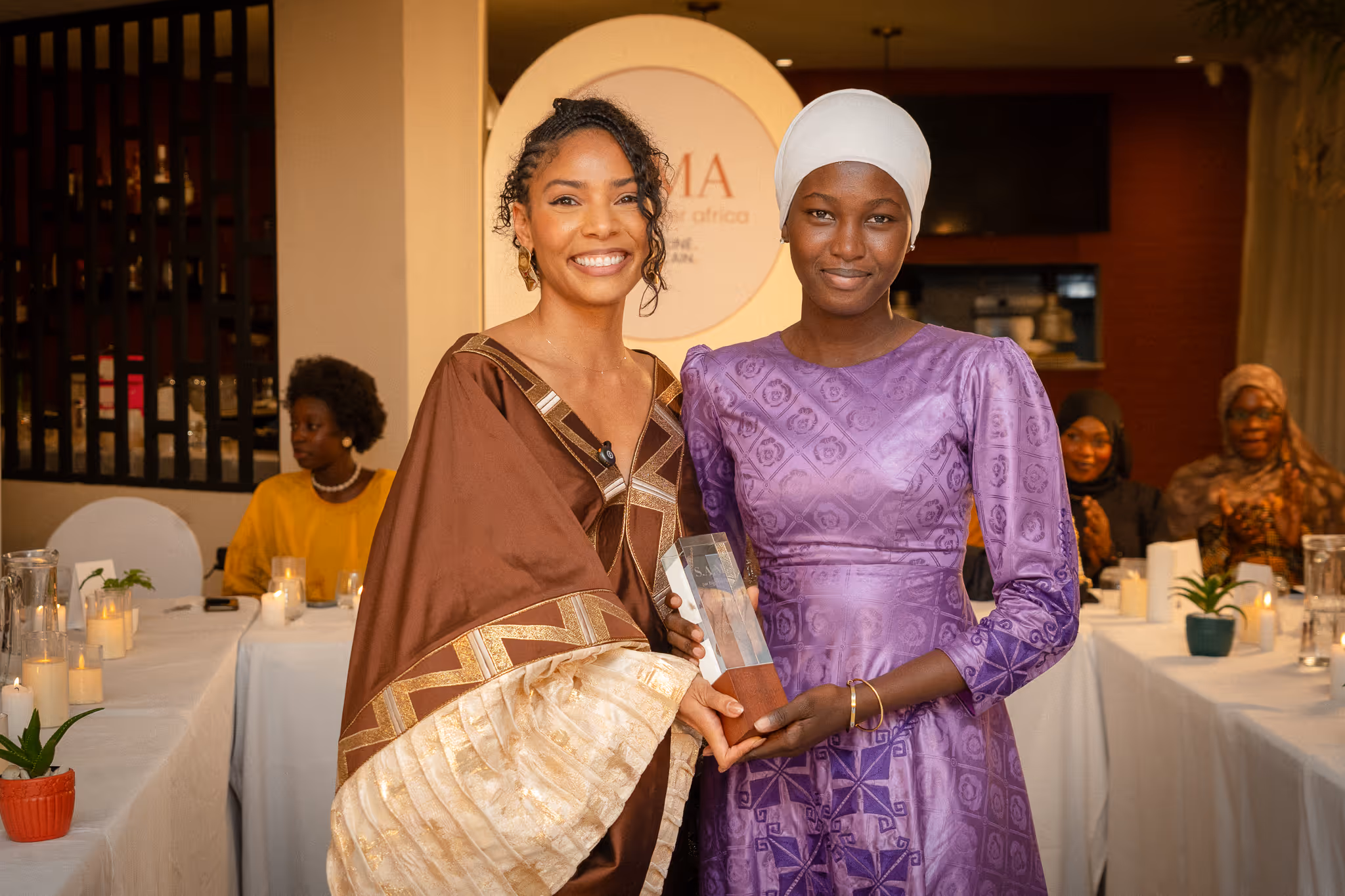 Two women smiling and holding an award together at a formal event with people applauding in the background.