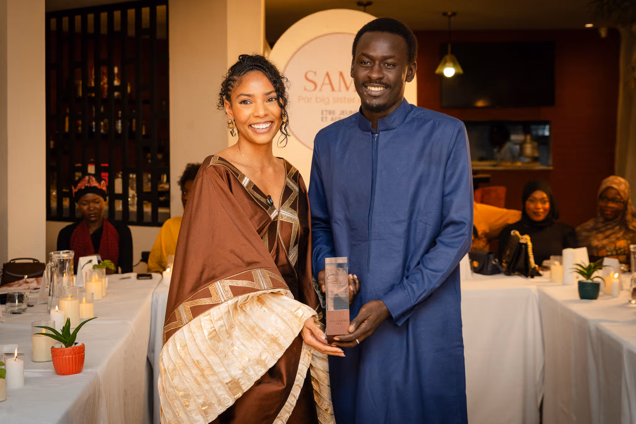Smiling man and woman in traditional attire holding an award together inside a warmly lit room with seated guests in the background.