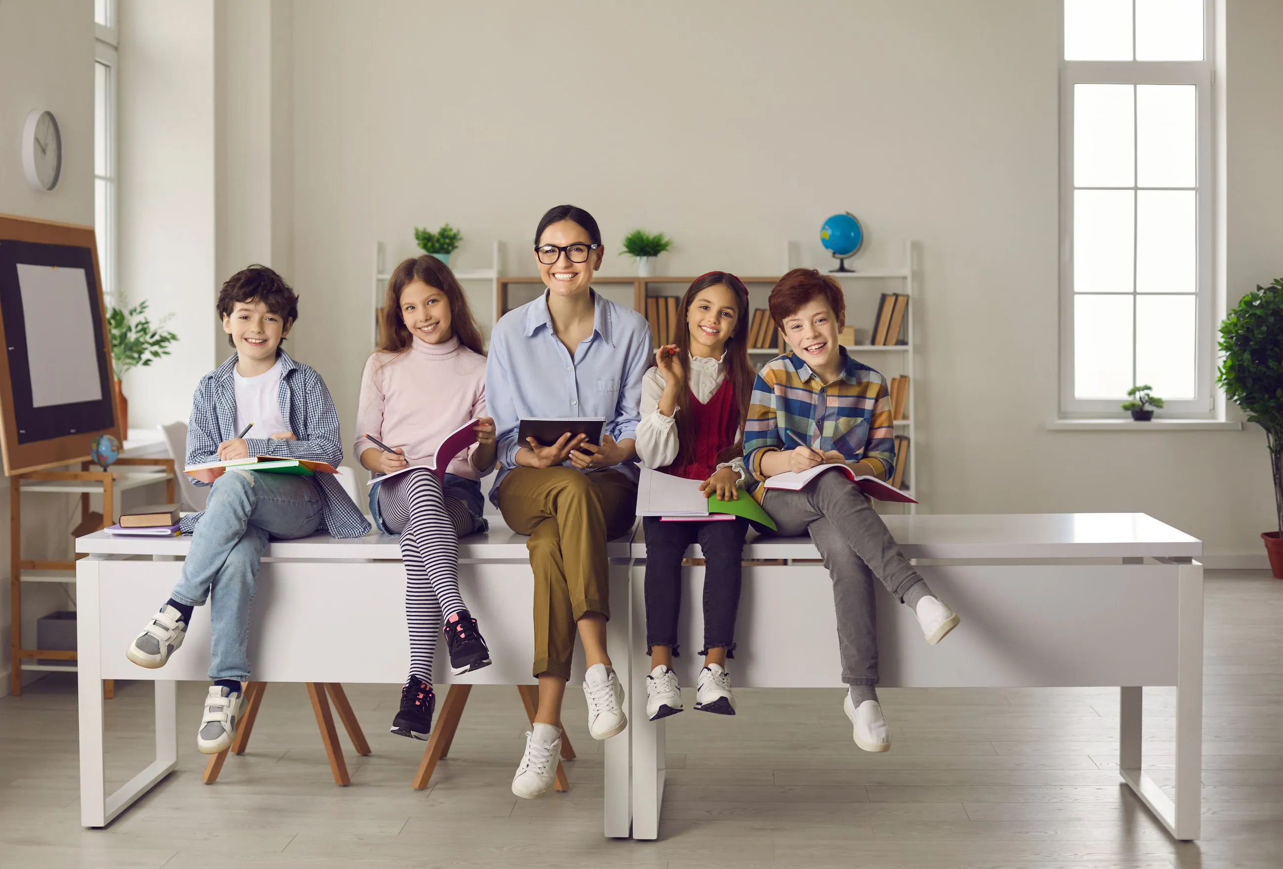 Nauczycielka siedząca z grupą uśmiechniętych dzieci w klasie, wszyscy trzymają książki.