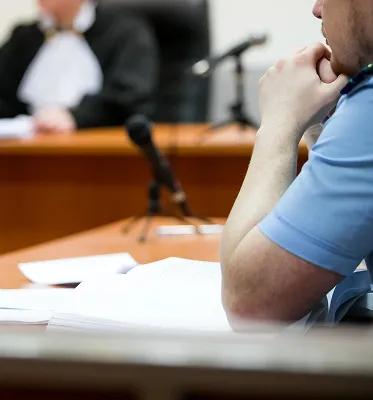 Close-up of a person in a blue shirt sitting at a desk with open documents, blurred figure in judicial robes in the background.