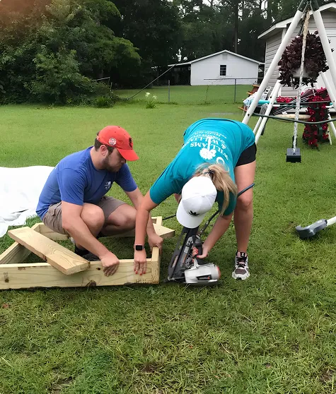 Two people working on assembling a wooden frame outdoors on grass, one holding the wood while the other uses a nail gun.