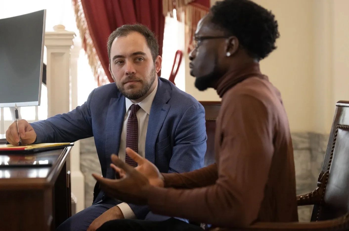 Two men having a serious conversation in an office, one wearing a blue suit and the other in a brown turtleneck.