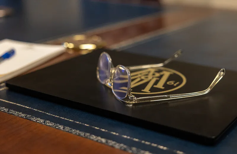 Transparent glasses resting on a black folder with a gold emblem on a blue and wooden desk.