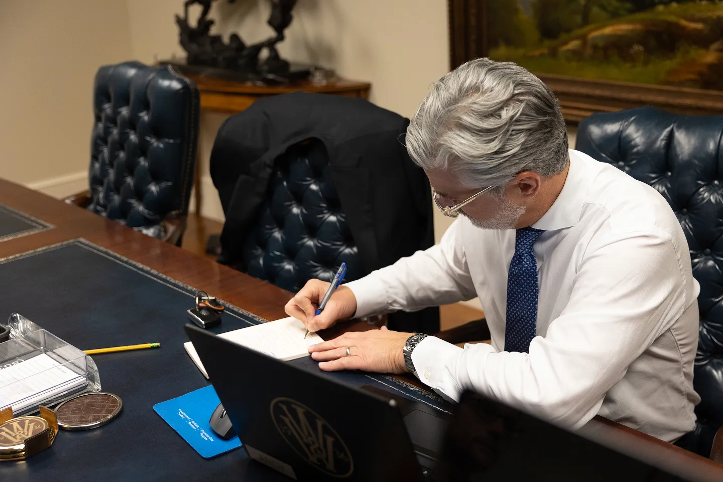 Man with gray hair and glasses in a white shirt and blue tie writing on a notepad at a desk with a laptop and office supplies.