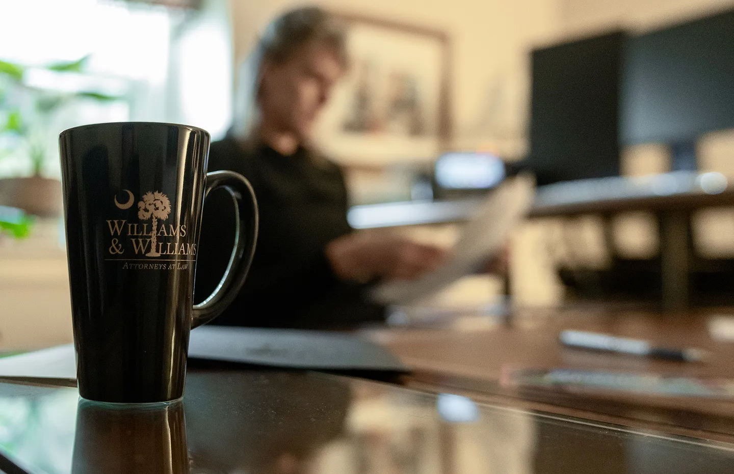 Black coffee mug with Williams & Williams Attorneys at Law logo on a desk, blurred person reading a document in the background.