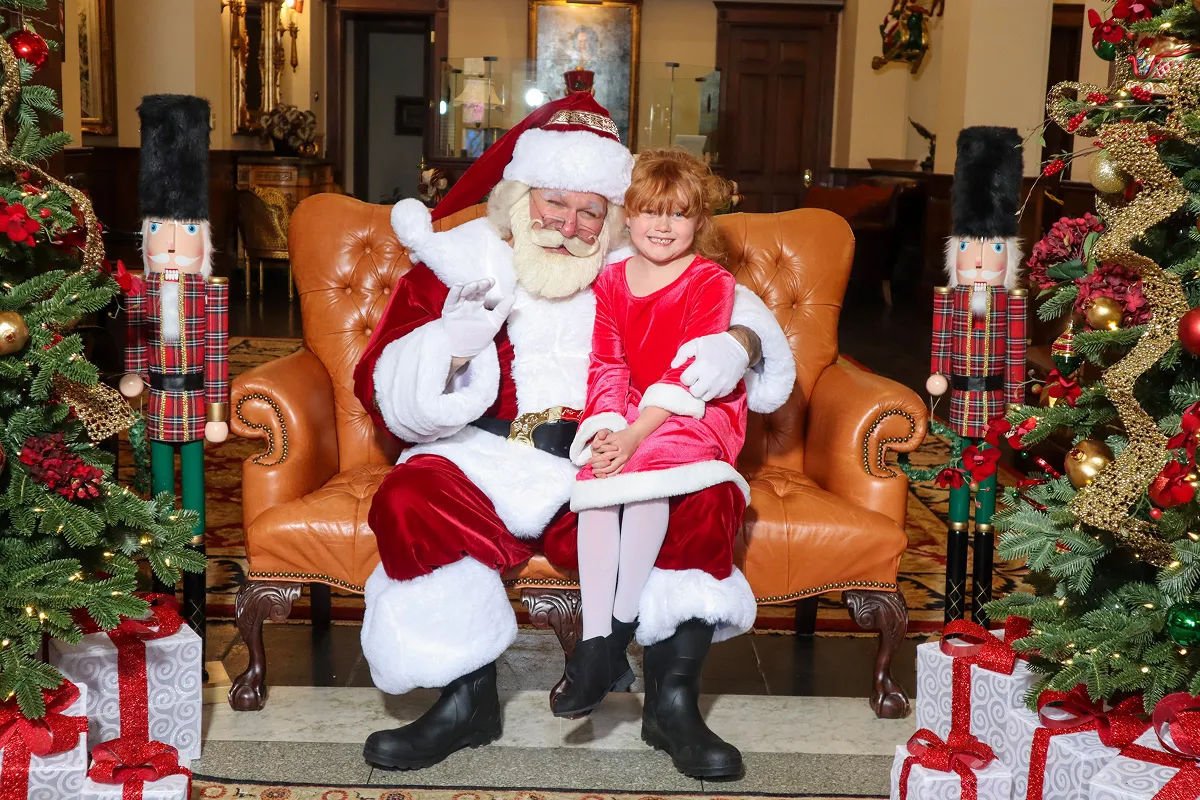 Santa Claus sitting on a brown leather chair holding a smiling young girl in a red dress, with Christmas trees, gift boxes, and nutcracker decorations on either side.