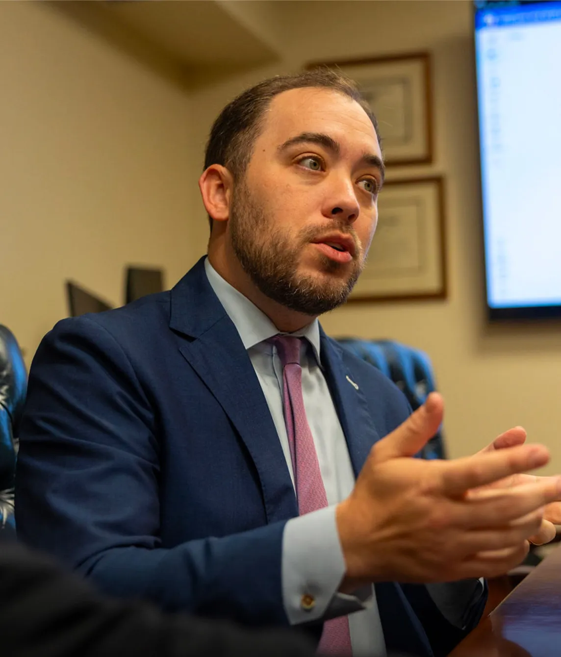 Man in blue suit and pink tie speaking and gesturing with his hands in an office setting.
