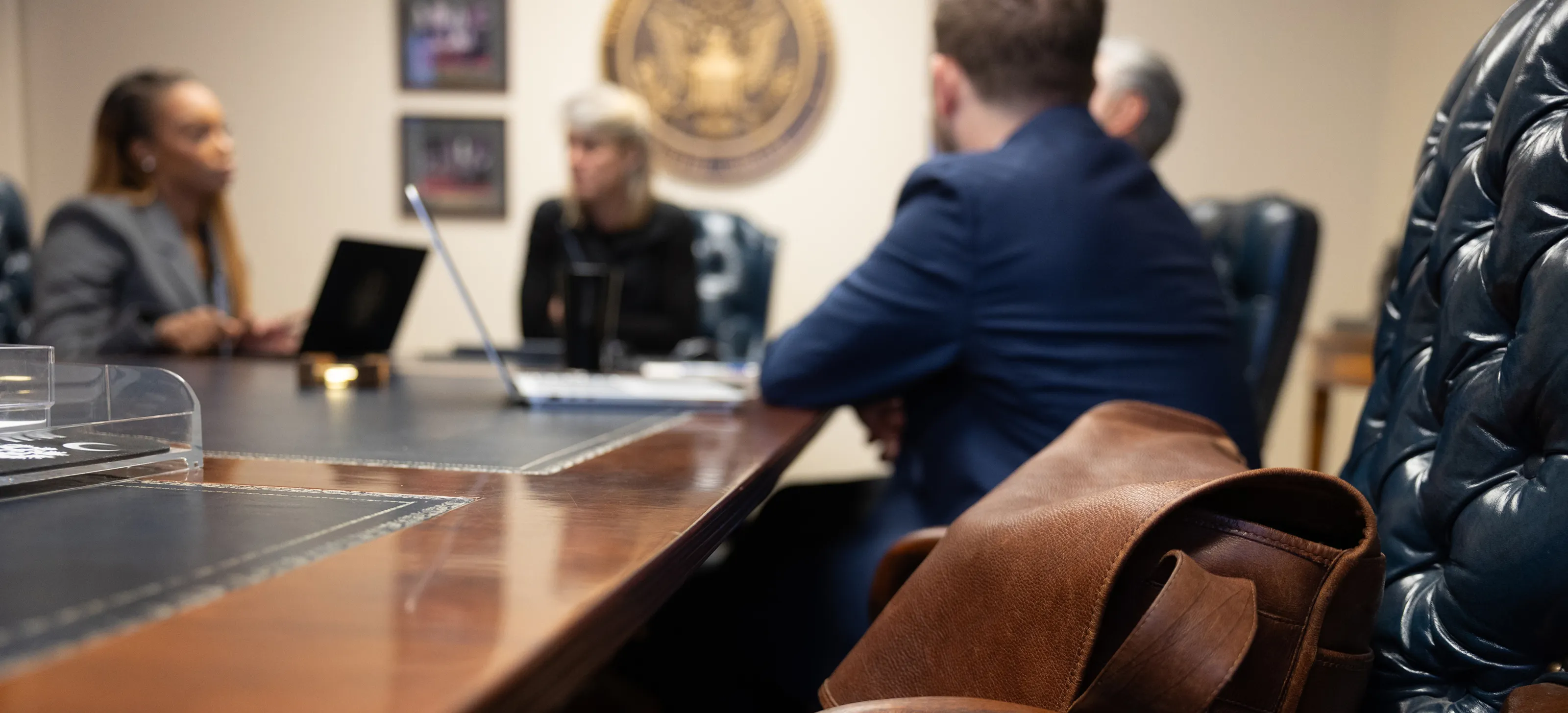 Blurred image of a business meeting with people sitting at a conference table and a laptop, with a brown leather bag in sharp focus in the foreground.