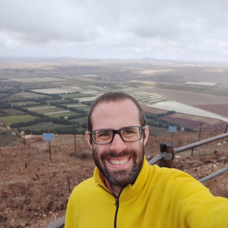 Smiling man with glasses holding a camera outdoors with a crowd and building in the background.