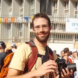 Smiling man with glasses holding a camera outdoors with a crowd and building in the background.