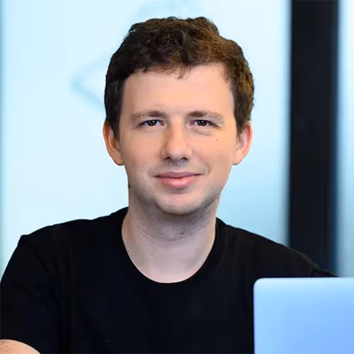 Young man with short curly brown hair wearing a black shirt, sitting indoors with a laptop in front of him.