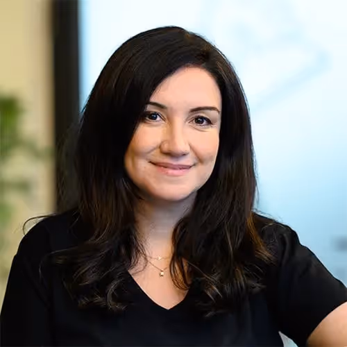 Smiling woman with long dark hair wearing a black shirt, seated indoors with a blurred background.