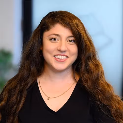Smiling woman with long curly brown hair wearing a black V-neck top and a thin gold necklace against a blurred indoor background.