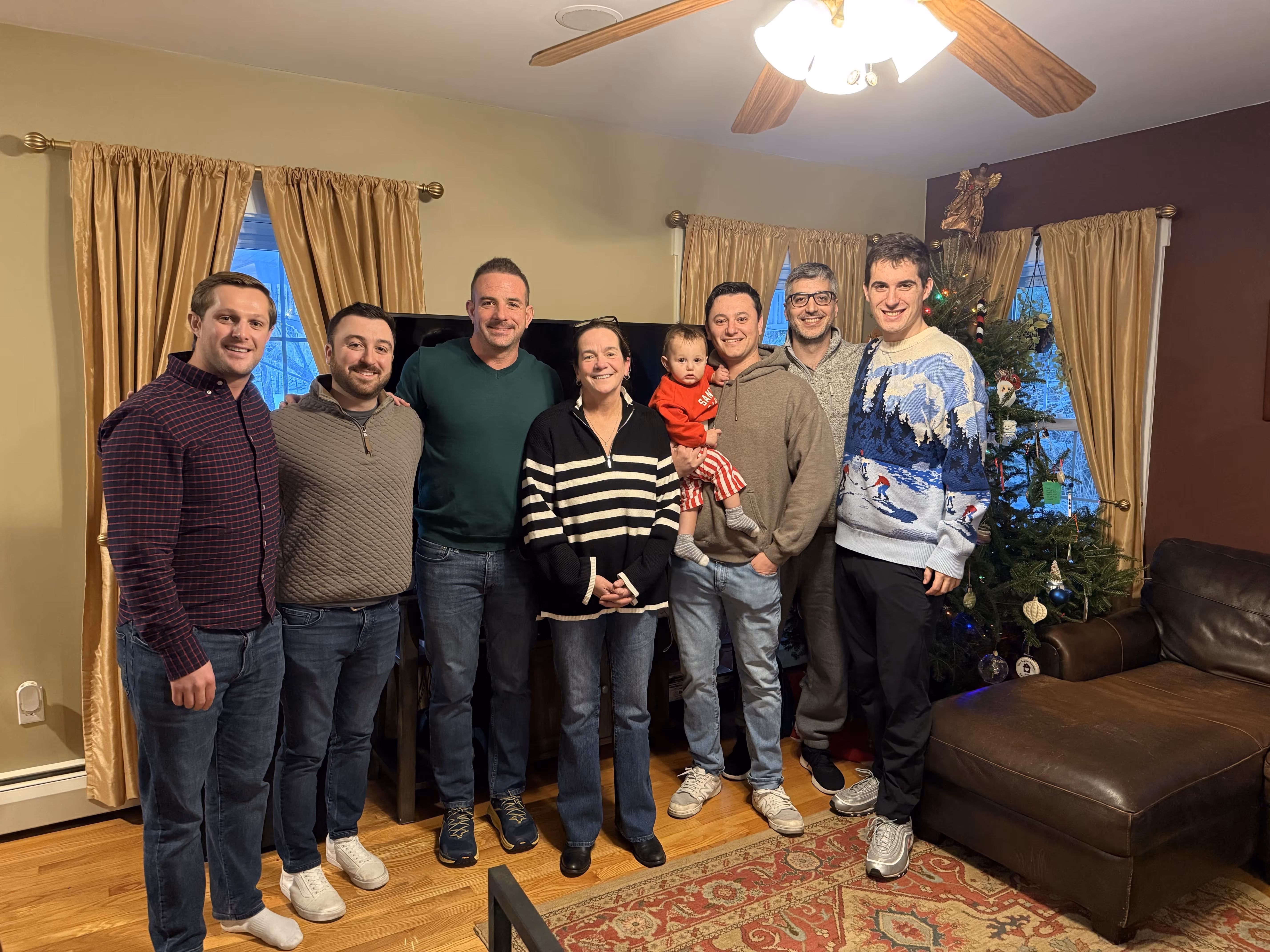 Seven adults and one child posing in a living room decorated with a Christmas tree and gold curtains.