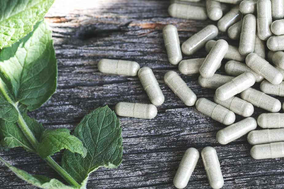 Green herbal leaves next to a pile of gray capsules on a wooden surface.