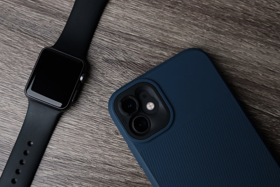 Black smartwatch with a square screen next to a smartphone in a dark blue textured case on a wooden surface.