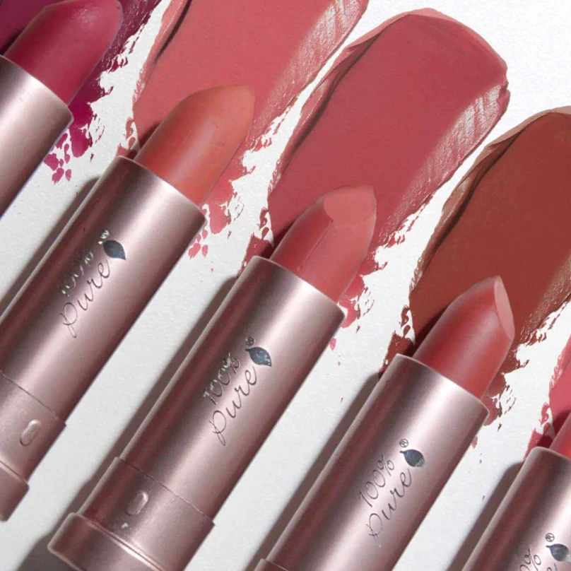 Close-up of five 100% Pure lipsticks in varying shades of pink and nude with matching swatches behind each.