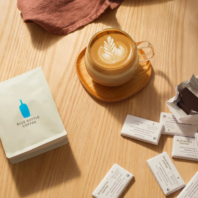 Glass cup of latte with leaf latte art on a wooden tray surrounded by Blue Bottle Coffee packaging and chocolate bars on a wooden table.