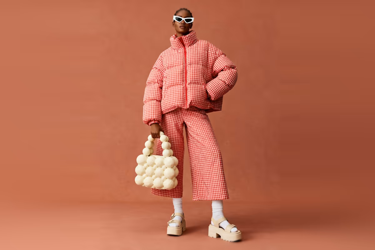 Model wearing a red and white checkered puffer jacket and matching wide-leg pants, holding a white bubble-style handbag, with white socks and chunky beige platform sandals against a brown background.