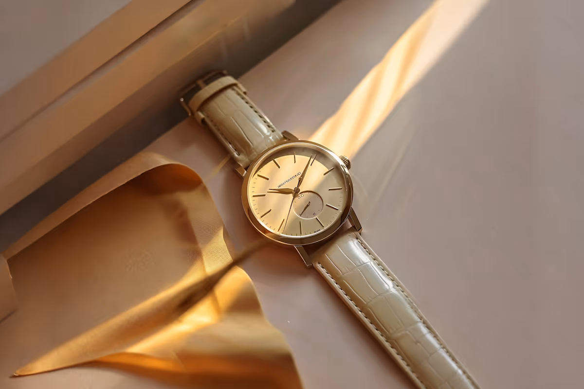 Elegant wristwatch with a light beige leather strap and gold-tone case resting on a smooth, glossy surface with warm sunlight casting shadows.