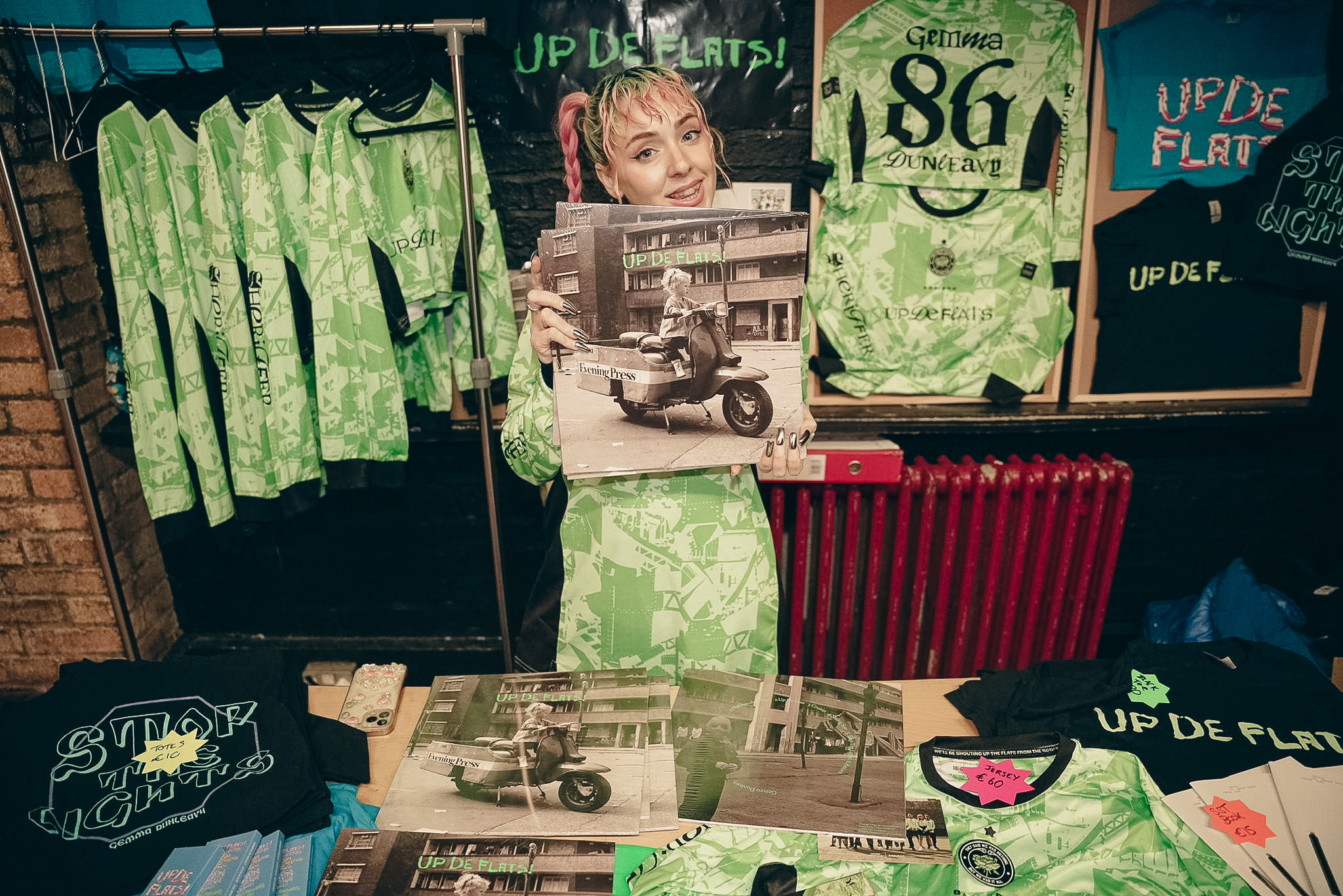 Person with pink hair holding vinyl records featuring a child on a scooter, standing behind a table displaying green patterned shirts, black t-shirts, and UP DE FLATS merchandise in a shop.