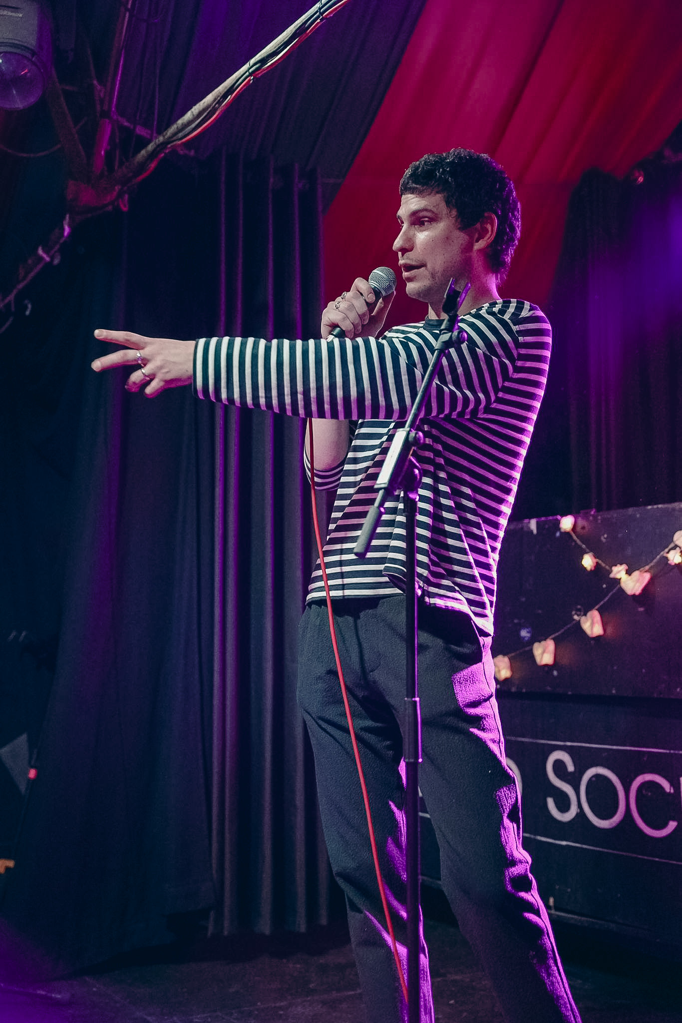 Man in black and white striped shirt speaking into a microphone on stage with a red and purple backdrop.