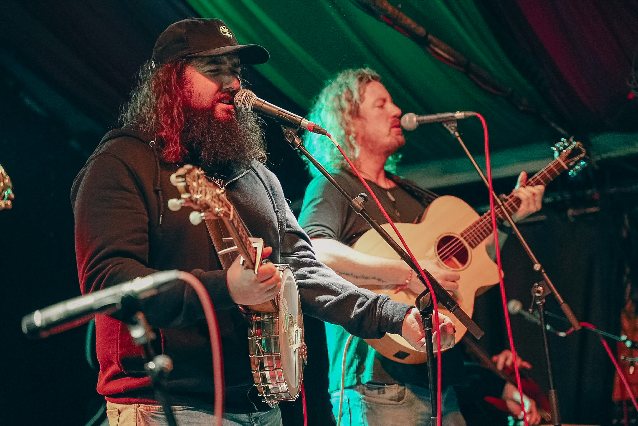 Two musicians performing on stage, one singing into a microphone while playing a banjo, the other playing an acoustic guitar.