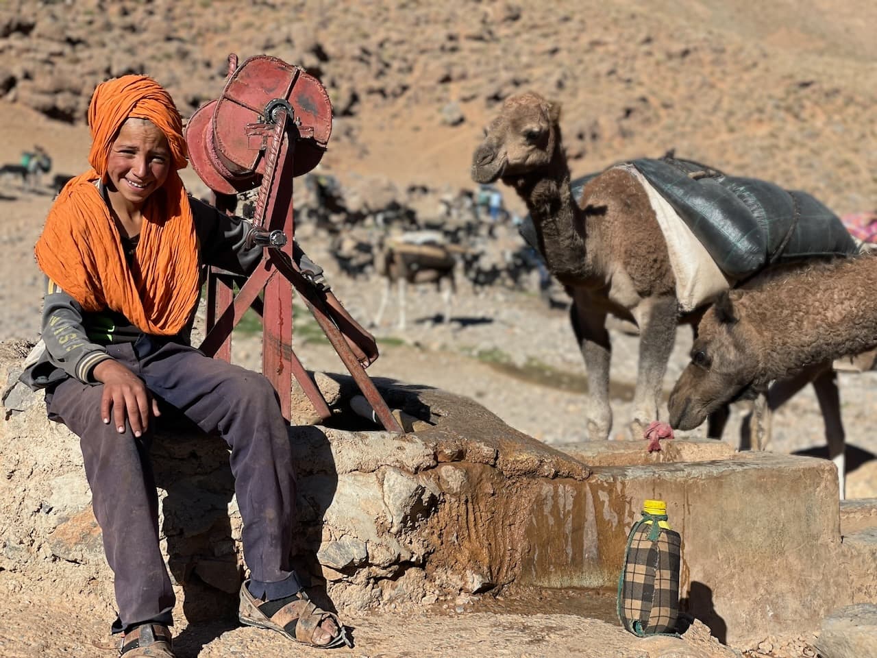 Portrait authentique d'un jeune garçon nomade de la tribu Aït Atta souriant, portant un chèche orange traditionnel, assis près d'un puits pastoral en pierre avec ses dromadaires dans un paysage de hamada rocailleuse du Sud marocain.