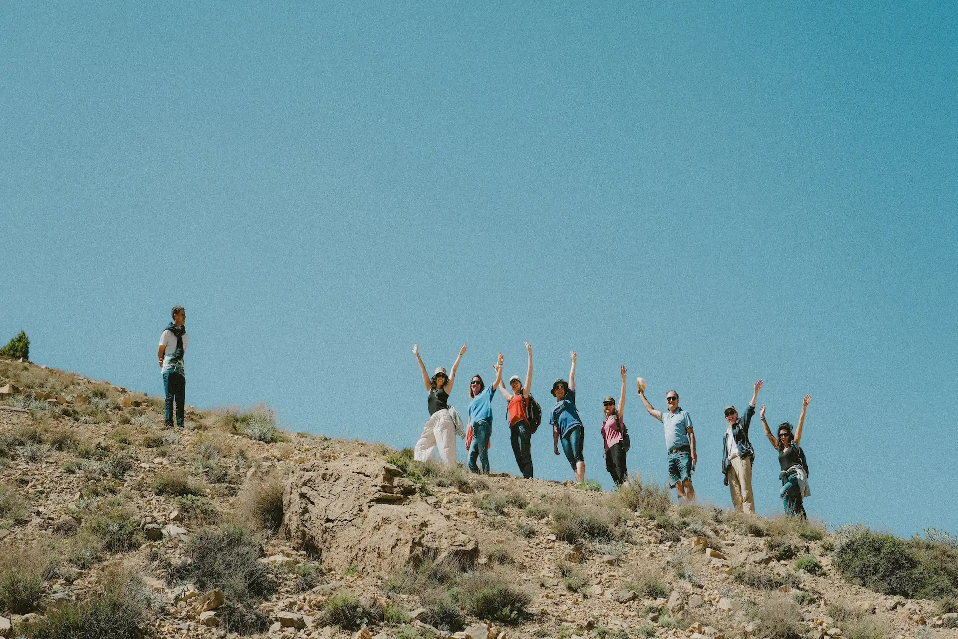 Un groupe de randonneurs debout sur une crête rocailleuse et aride, les bras levés vers le ciel bleu, célébrant leur ascension lors d'une excursion en plein air.