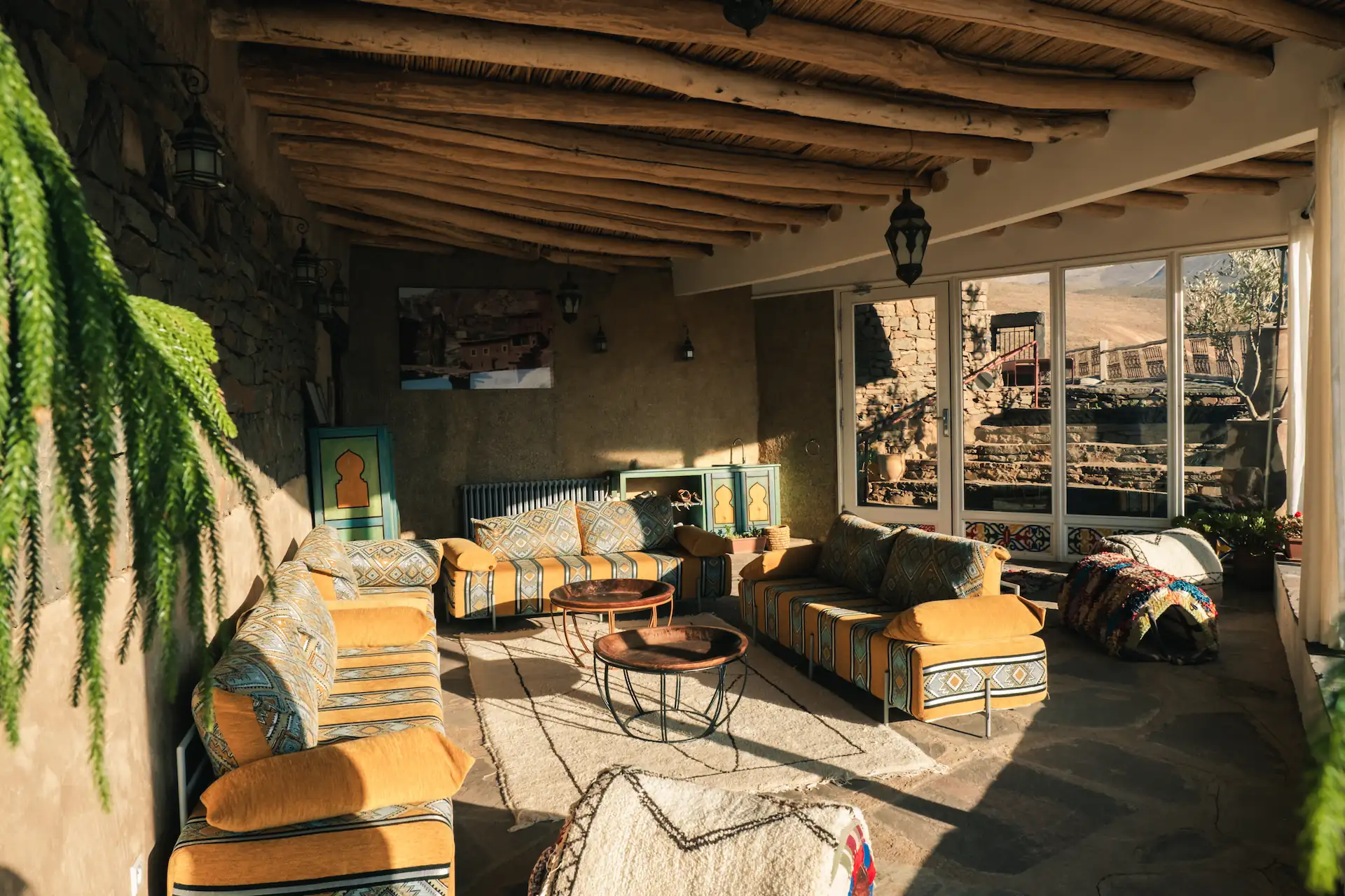 Außenlounge einer marokkanischen Ecolodge mit gelben und blauen Berber-Sofas, handgewebten Teppichen, Kupfertischen und Blick auf das Atlasgebirge durch große Glastüren.