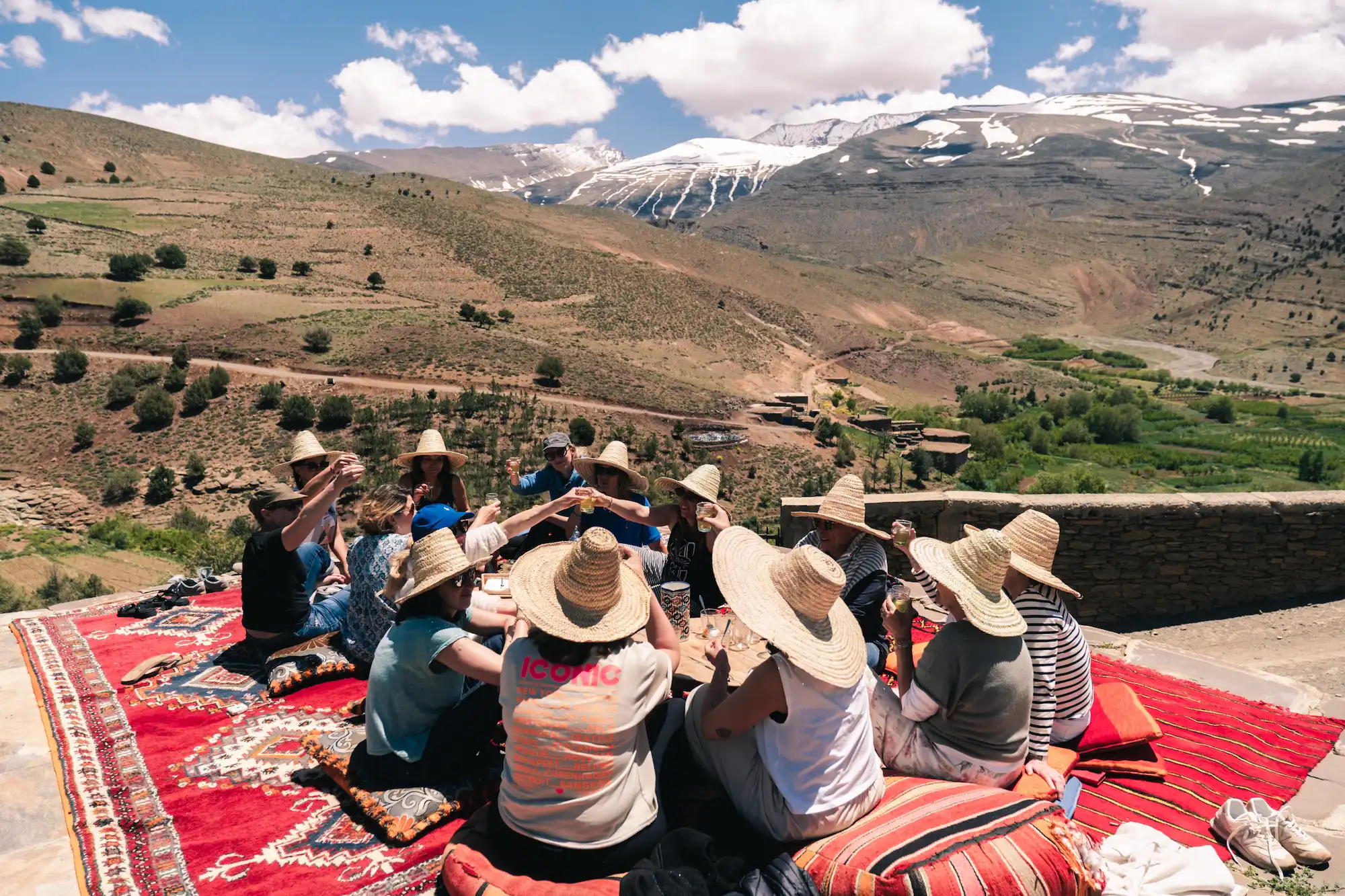 Groupe de voyageurs portant des chapeaux de paille, assis sur des tapis berbères rouges, trinquant face aux montagnes enneigées de l'Atlas marocain.
