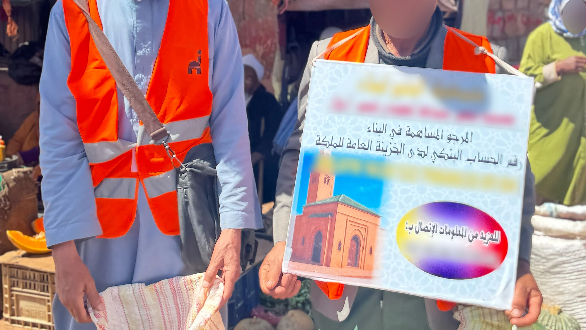 Deux collecteurs de dons bénévoles en gilets orange tiennent un panneau d'appel aux dons pour la construction d'une mosquée dans un souk rural marocain.