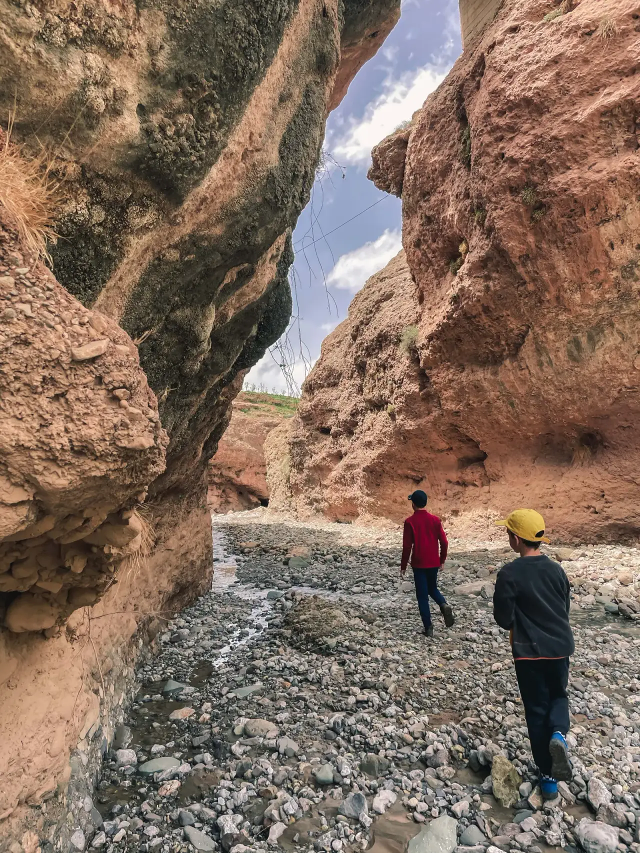 Children hiking along a dry riverbed between towering red canyon cliffs with Touda Ecolodge.